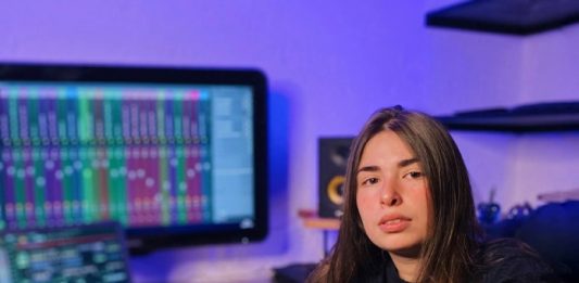 dj katrip posa para a foto em seu escritório, em frente a uma grande tela de computador; ela está com um braço sobre uma das pernas em cima da cadeira