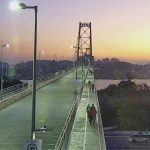 camera de monitoramento sobre cabeceira insular da ponte em por do sol; pessoas andando na passarela; poste no meio da imagem