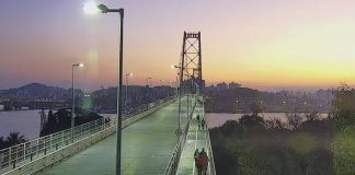 camera de monitoramento sobre cabeceira insular da ponte em por do sol; pessoas andando na passarela; poste no meio da imagem