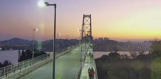 camera de monitoramento sobre cabeceira insular da ponte em por do sol; pessoas andando na passarela; poste no meio da imagem