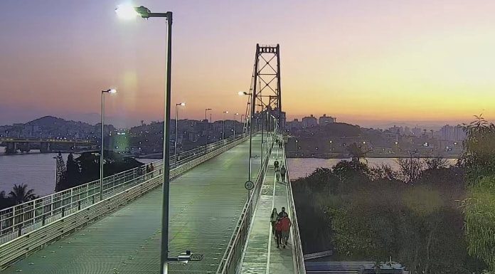 camera de monitoramento sobre cabeceira insular da ponte em por do sol; pessoas andando na passarela; poste no meio da imagem