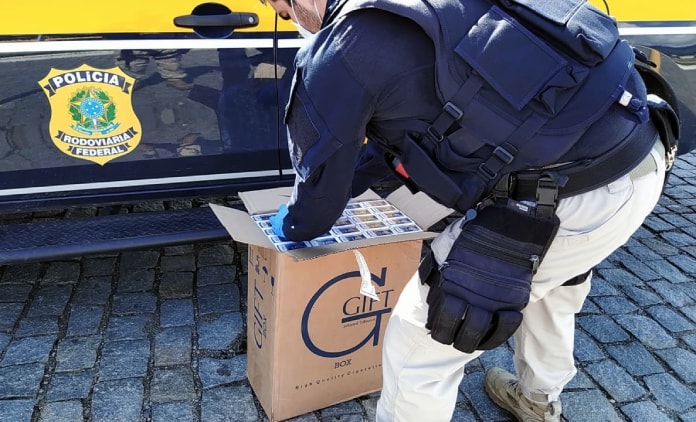 policial se abaixa para abrir caixa de cigarros gift no chão ao lado da viatura da PRF