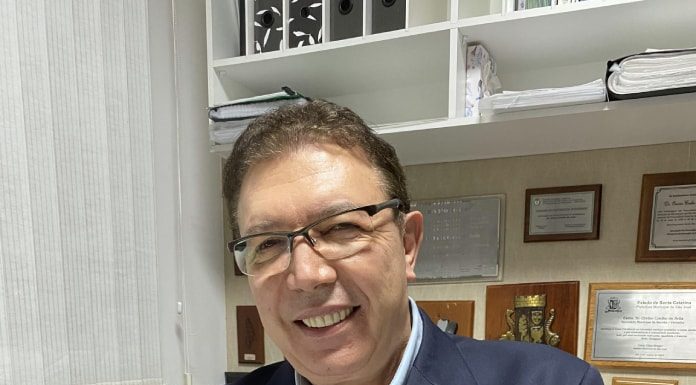 orvino em escritório olha sorridente para a câmera; ele usa terno e óculos. quadros ao fundo