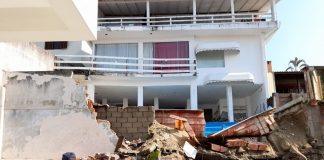 muro desabado com escombros sobre terreno vizinho; casa de três andares ao fundo; dois bombeiros na frente