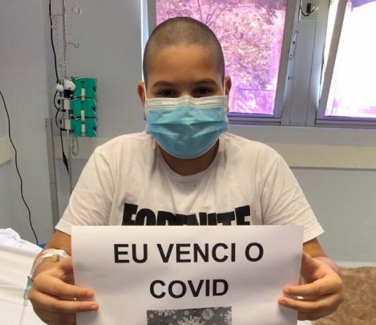 Garoto de 10 anos recebe alta após 4 dias na UTI com Covid-19 garoto usando máscara sentado na beira da cama do hospital mostra para foto papel com inscrição "eu venci o covid"