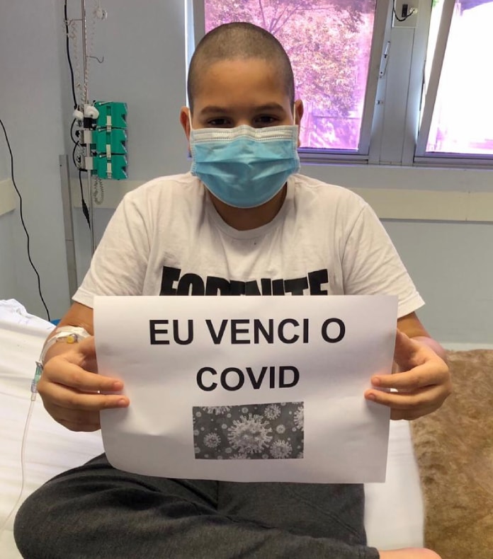 garoto usando máscara sentado na beira da cama do hospital mostra para foto papel com inscrição "eu venci o covid"