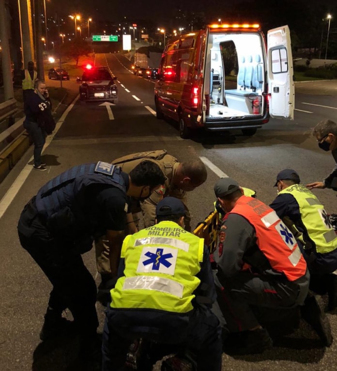 socorristas e policias abaixados em volta do motociclista, que não aparece; ao fundo ambulância e viatura sobre ponte