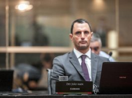 deputado joão amin na bancada da alesc olha para o alto e há uma placa com seu nome à frente junto com notebook