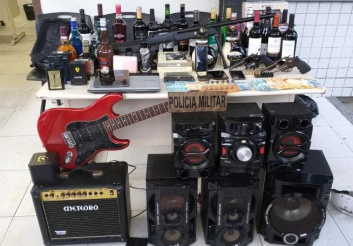 diversos produtos como caixas de som, guitarra, bebidas e eletrônicos organizados sobre uma mesa com placa da pm por cima