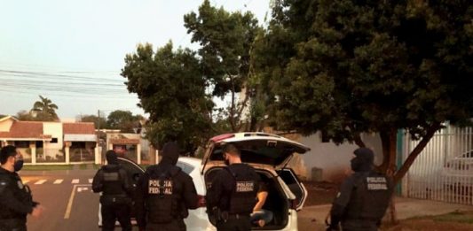 policias uniformizados atrás de um carro com porta malas aberto estacionado em área residencial com casas ao fundo e árvores na calçada