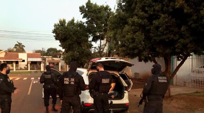 policias uniformizados atrás de um carro com porta malas aberto estacionado em área residencial com casas ao fundo e árvores na calçada