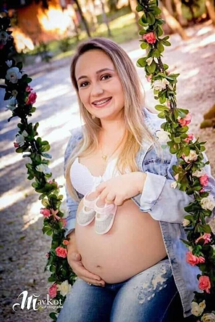 flavia godinho mafra em foto grávida sentada em balanço com flores segura um sapatinho de bebê sobre a barriga