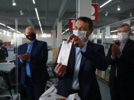 moisés segura com as duas mãos uma nota fiscal; ele usa máscara; ao fundo outros homens de máscara ao redor de caixa de uma grande loja
