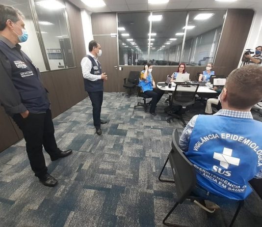 motta e moisés em pé conversando com servidoras com coletes do governo estadual em diferentes mesas em uma sala acarpetada; todos usam máscaras