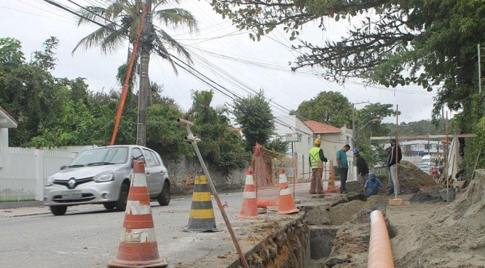 vala aberta no canto da rua, com cano dentro, operários e cones em volta; carro passando ao lado na rua