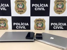 dois notebooks e dois celulares sobre mesa com painel da polícia civil ao fundo