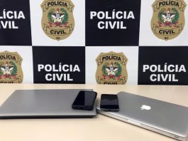 Polícia investiga tentativa de extorsão cibernética contra empresa de vigilância de São José dois notebooks e dois celulares sobre mesa com painel da polícia civil ao fundo