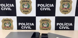 dois notebooks e dois celulares sobre mesa com painel da polícia civil ao fundo