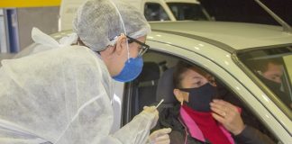 profissional de saúde trajada com epi se inclina para janela de carro onde está mulher e homem com máscaras dentro do veículo