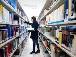mulher com casaco preto, em frente uma prateleira de biblioteca, segurando um livro na mão