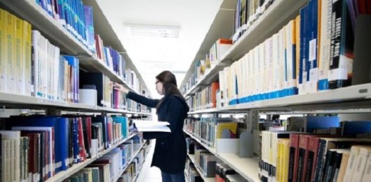 mulher com casaco preto, em frente uma prateleira de biblioteca, segurando um livro na mão