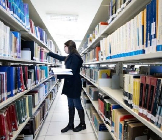 Capes publica novas regras para concessão de bolsas de pós-graduação mulher com casaco preto, em frente uma prateleira de biblioteca, segurando um livro na mão
