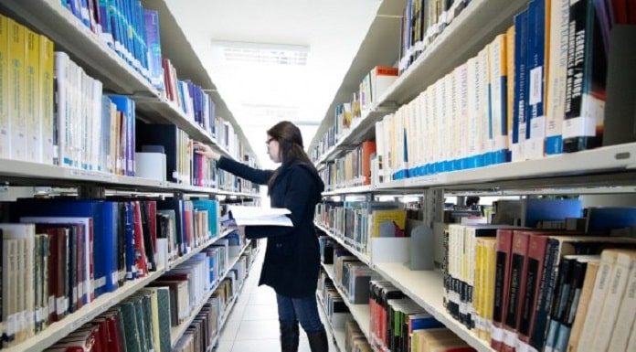 mulher com casaco preto, em frente uma prateleira de biblioteca, segurando um livro na mão