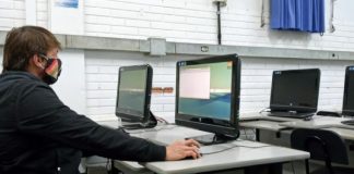 homem usando máscara sentado em frente a um computador em sala com mais computadores