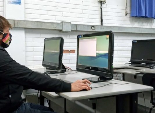 homem usando máscara sentado em frente a um computador em sala com mais computadores