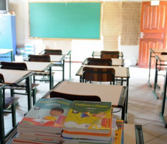 Definições para volta das atividades pedagógicas presenciais no estado livros didáticos sobre uma carteira em sala de aula com outras mesas e carteiras organizadas; porta aberta ao lado do quadro