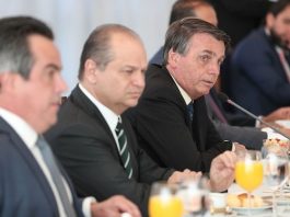Auxílio emergencial será prorrogado até o fim do ano homens sentados em uma mesa de reuniões, com copos de suco de laranja a sua frente, escutando o presidente Bolsonaro falar