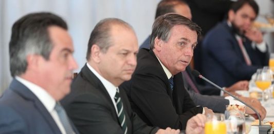 Auxílio emergencial será prorrogado até o fim do ano homens sentados em uma mesa de reuniões, com copos de suco de laranja a sua frente, escutando o presidente Bolsonaro falar