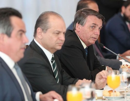 homens sentados em uma mesa de reuniões, com copos de suco de laranja a sua frente, escutando o presidente Bolsonaro falar