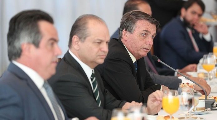 homens sentados em uma mesa de reuniões, com copos de suco de laranja a sua frente, escutando o presidente Bolsonaro falar