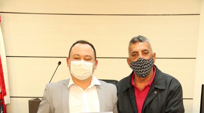 homem usando máscara e jaqueta preta entregando placa comemorativa para policial militar usando terno cinza