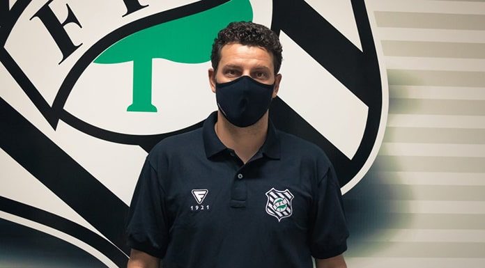 Homem vestindo camisa preta, usando máscara preta de pé em frente ao simbolo do Figueirense