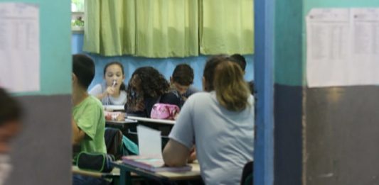 alunos vistos sentados nas carteiras de sala de aula por entre a porta