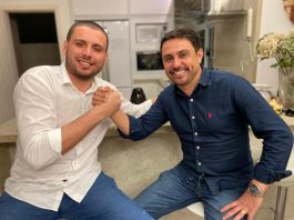 helio costa jr e fernando anselmo de mãos dadas sorridentes para a foto; eles estão sentado lado a lado em bancos altos