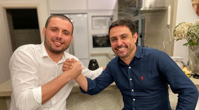 helio costa jr e fernando anselmo de mãos dadas sorridentes para a foto; eles estão sentado lado a lado em bancos altos