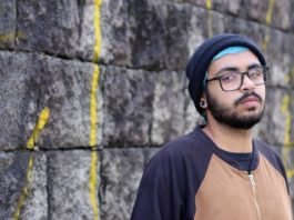 rapper warllock posa para a foto em frente a um muro; ele usa touca, óculos e tem barba