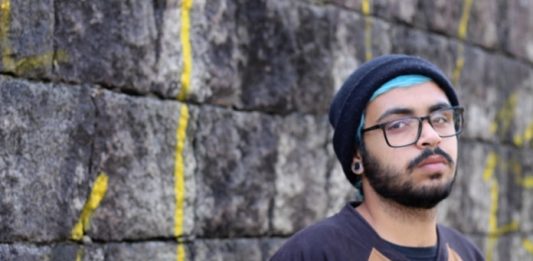 rapper warllock posa para a foto em frente a um muro; ele usa touca, óculos e tem barba