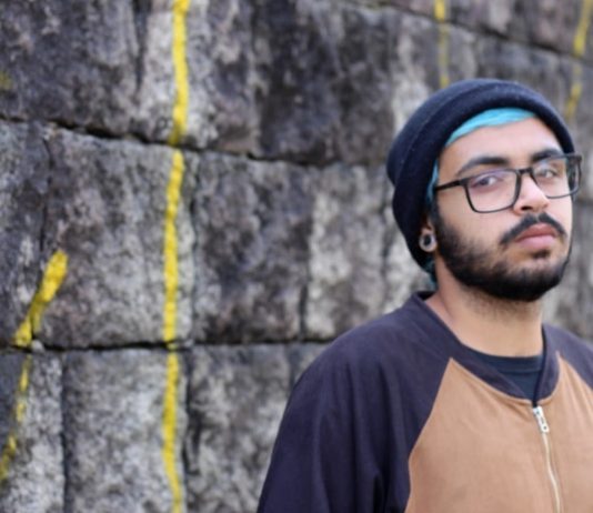 rapper warllock posa para a foto em frente a um muro; ele usa touca, óculos e tem barba