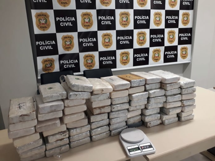 dezenas de pacotes empilhados sobre mesa com balança na frente e painel da polícia civil ao fundo