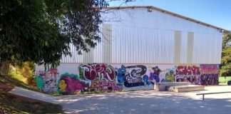 área de pista de skate com galpão ao fundo com desenhos de grafite no muro; área sombreada por árvores
