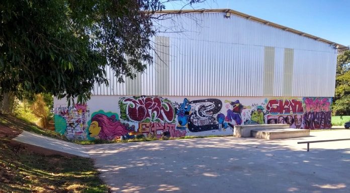 área de pista de skate com galpão ao fundo com desenhos de grafite no muro; área sombreada por árvores