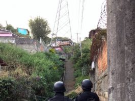 dois policiais do bope uniformizados vistos de costas em área do morro; torres de transmissão no alto