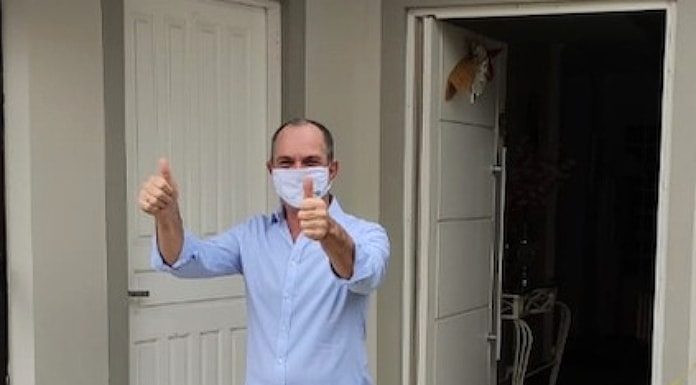 moacir usando máscara faz sinal de positivo com as duas mãos pra frente em frente à porta aberta de sua casa; ele usa camisa azul, calça jeans e chinelos
