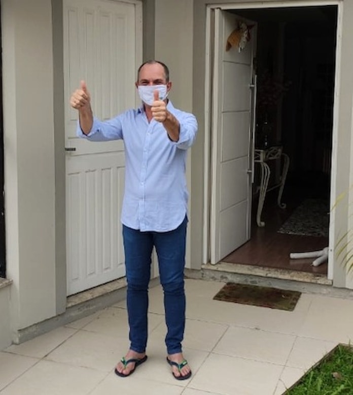 moacir usando máscara faz sinal de positivo com as duas mãos pra frente em frente à porta aberta de sua casa; ele usa camisa azul, calça jeans e chinelos