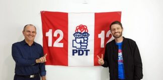 moacir da silva e andre guesser posam sorridentes para a foto em frente a uma bandeira do pdt; moacir faz o numero 1 com a mão e andré o dois