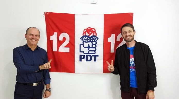 moacir da silva e andre guesser posam sorridentes para a foto em frente a uma bandeira do pdt; moacir faz o numero 1 com a mão e andré o dois
